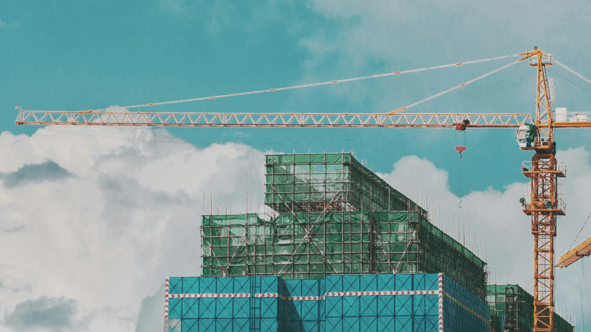 A yellow tower crane overseeing buildings under construction, covered in green netting, against a blue sky with clouds.